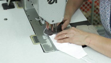 a seamstress sewing the strip of elastic garter making ear loops on a white face mask in a sew factory - covid-19 pandemic - medium shot pan right