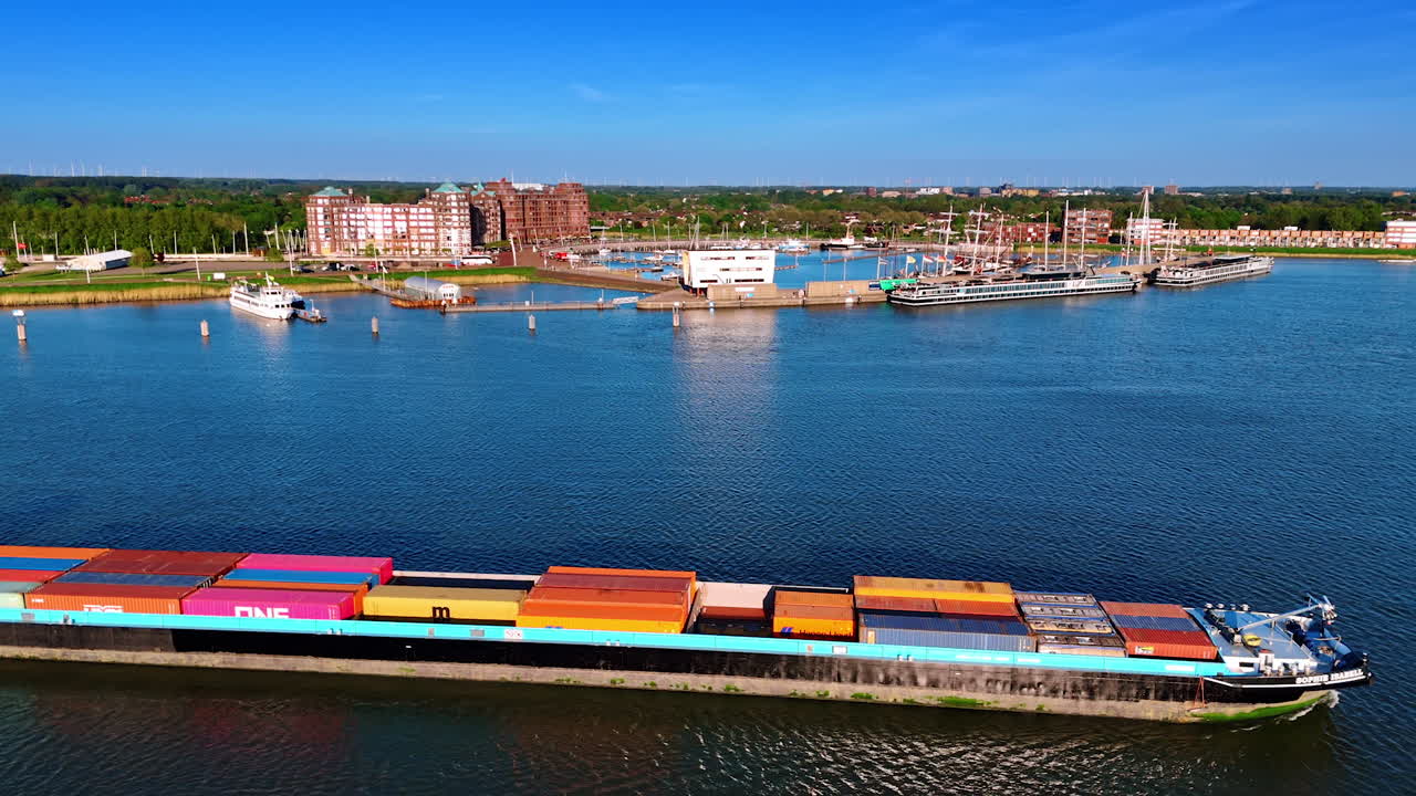 Barge loaded with colorful containers moves by the lake Markermeer. Port of Lelystad, the Netherlands at backdrop. Aerial view.