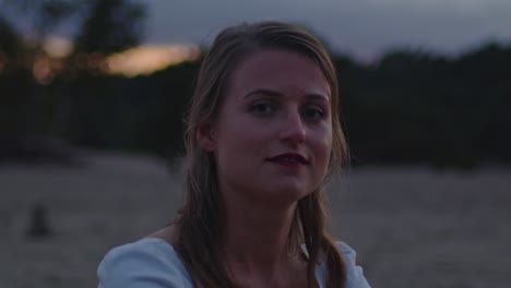 close up of young woman sitting in sand dunes at dusk and looking at camera