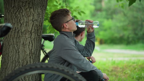 two young siblings take a break under a tree, one drinks water while the other looks into the distance holding a water bottle, the background features a blur of greenery and natural surroundings