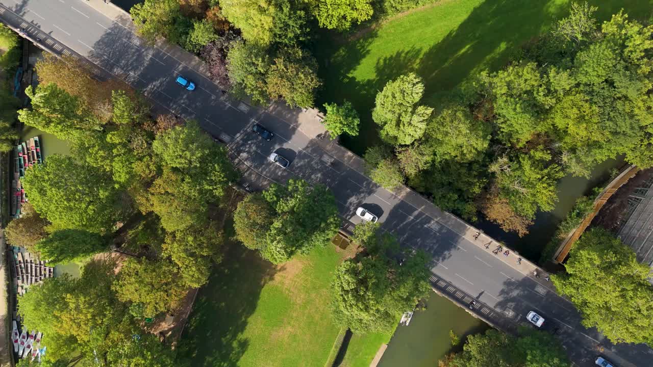 Aerial top-down drone shot at 60m height. View rotates anticlockwise over a two-lane road as vehicles and pedestrians enter the frame. Trees, grass, and pathways line both sides during a full orbit