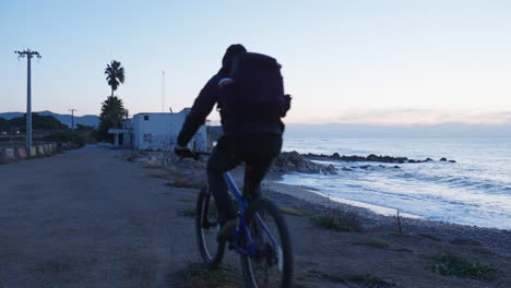 a traveler rides his bike towards a beach house, coming to a stop at the edge of the rocky shore to take in the view of the ocean during the blue hour