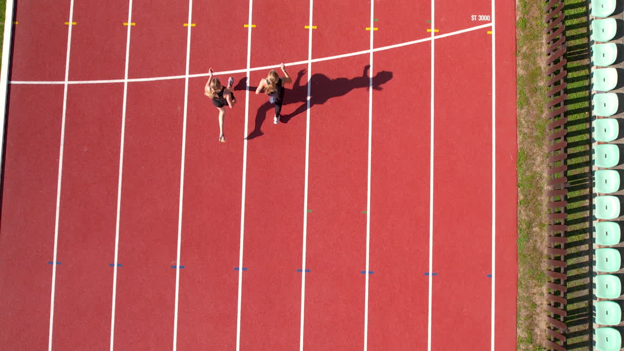 Aerial shot of two athletes running in sync along a curved section of a red track, shadows extending to the side, captured from above