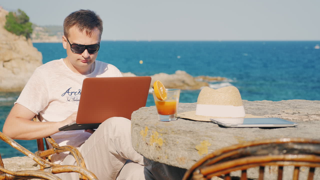 hombre con gafas de sol trabaja con una laptop contra el fondo del mar en una mesa en un café