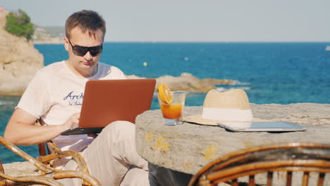 hombre con gafas de sol trabaja con una laptop contra el fondo del mar en una mesa en un café