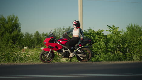 una motociclista femenina se sienta relajada en su bicicleta deportiva roja con su chaqueta abierta, mirando hacia atrás mientras los coches pasan por la carretera