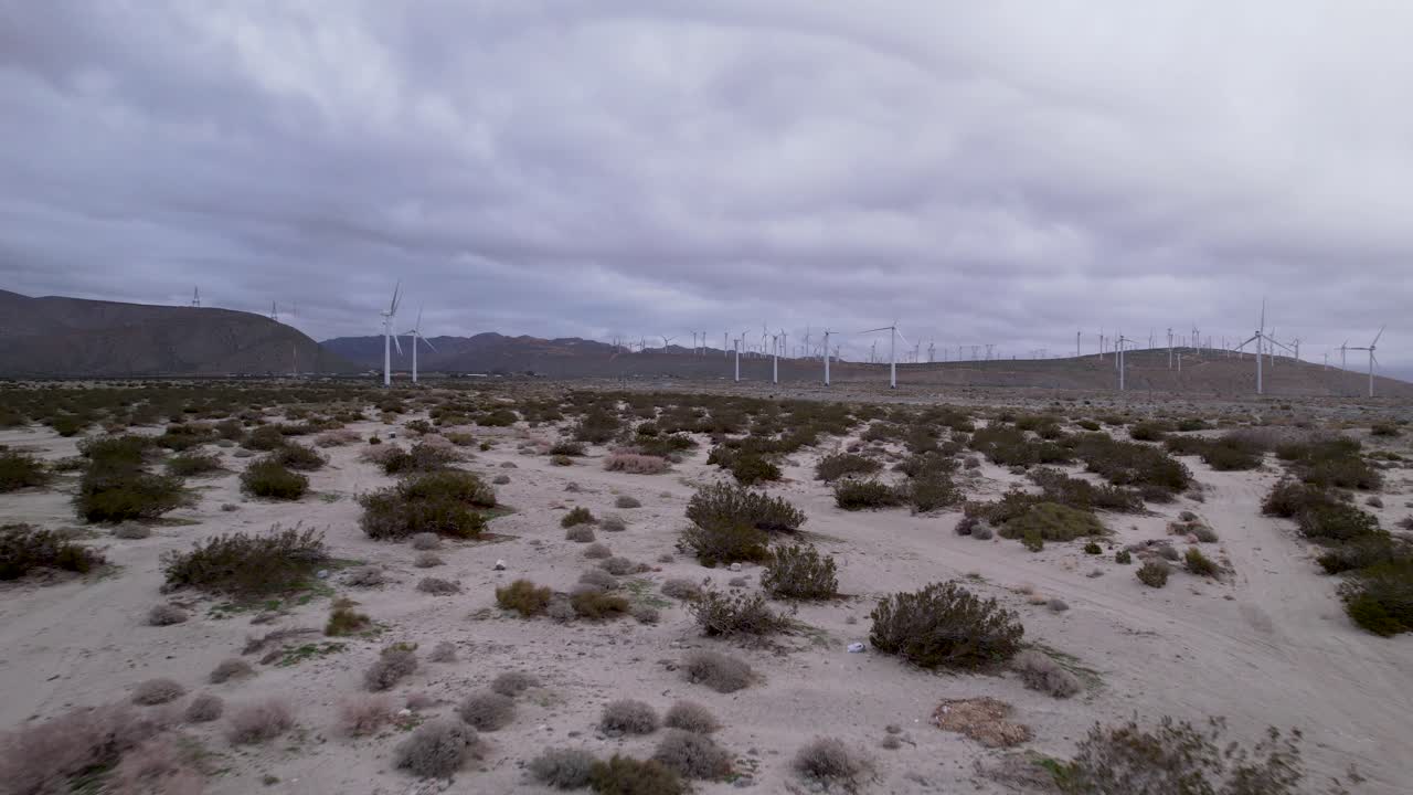 Carrellata aerea lenta di un parco eolico nel deserto di Palm Springs in una giornata nuvolosa con montagne sullo sfondo