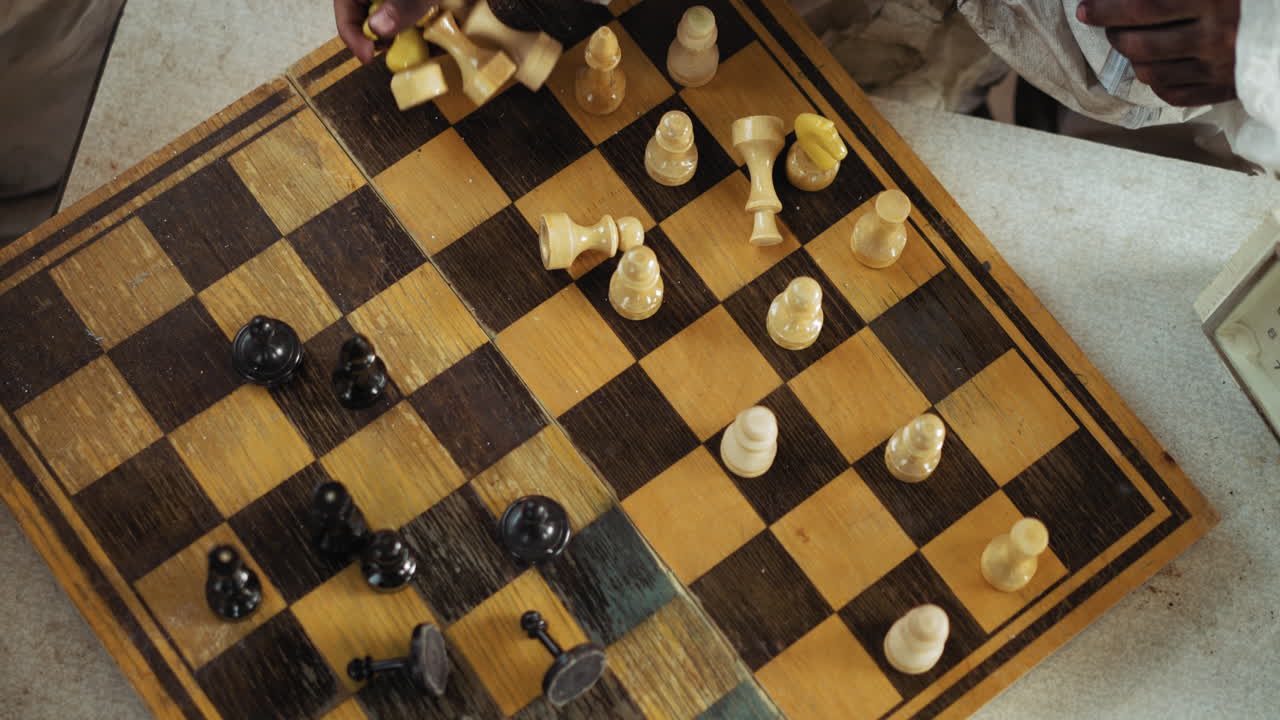 Close up top view of person placing chess pieces on worn wooden chessboard during intense match, with scattered black and white pieces suggesting strategic thinking, and competitive interaction
