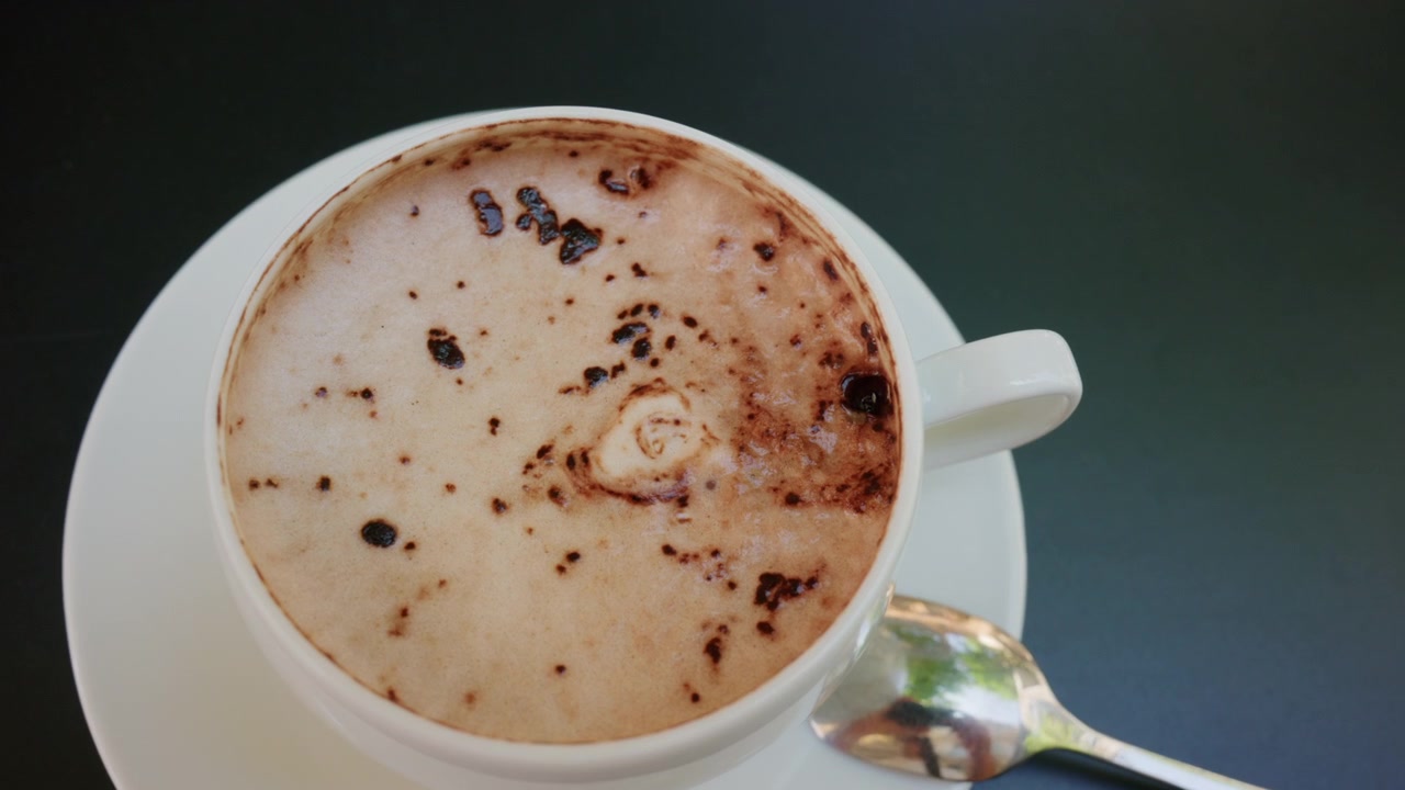 Cappuccino chaud avec une mousse onctueuse et des vermicelles de chocolat, servi dans une tasse blanche avec une cuillère sur une table sombre.