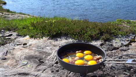 egg and bacon cooked on gas fired camping frying pan out in nature with a fresh mountain lake seen in background - norway europe