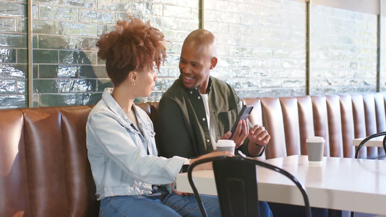 African American couple sitting in booth, man showing smartphone, woman tapping screen, coffee cups
