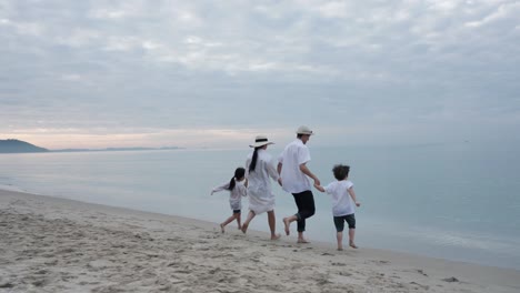 side view happy asian family on summer vacation father, mother, son and daughter running hand in hand together on the beach in the morning time, sunrise. holiday and travel concept. slow motion