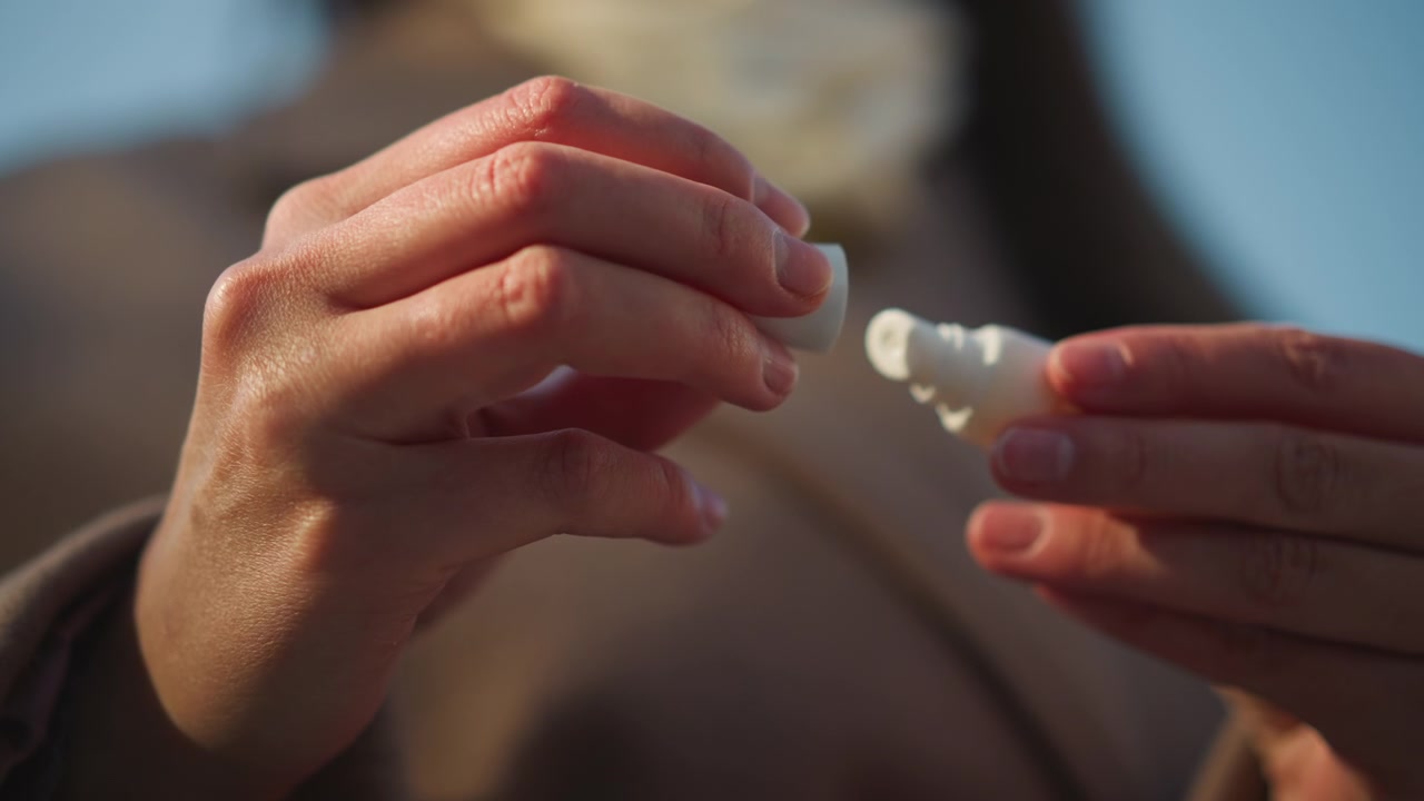 Primo piano di mani che aprono un piccolo tubetto bianco rivelandone il contenuto. La scena si concentra sull'interazione tattile con la confezione di un prodotto per la cura personale su uno sfondo esterno con luce naturale soffusa