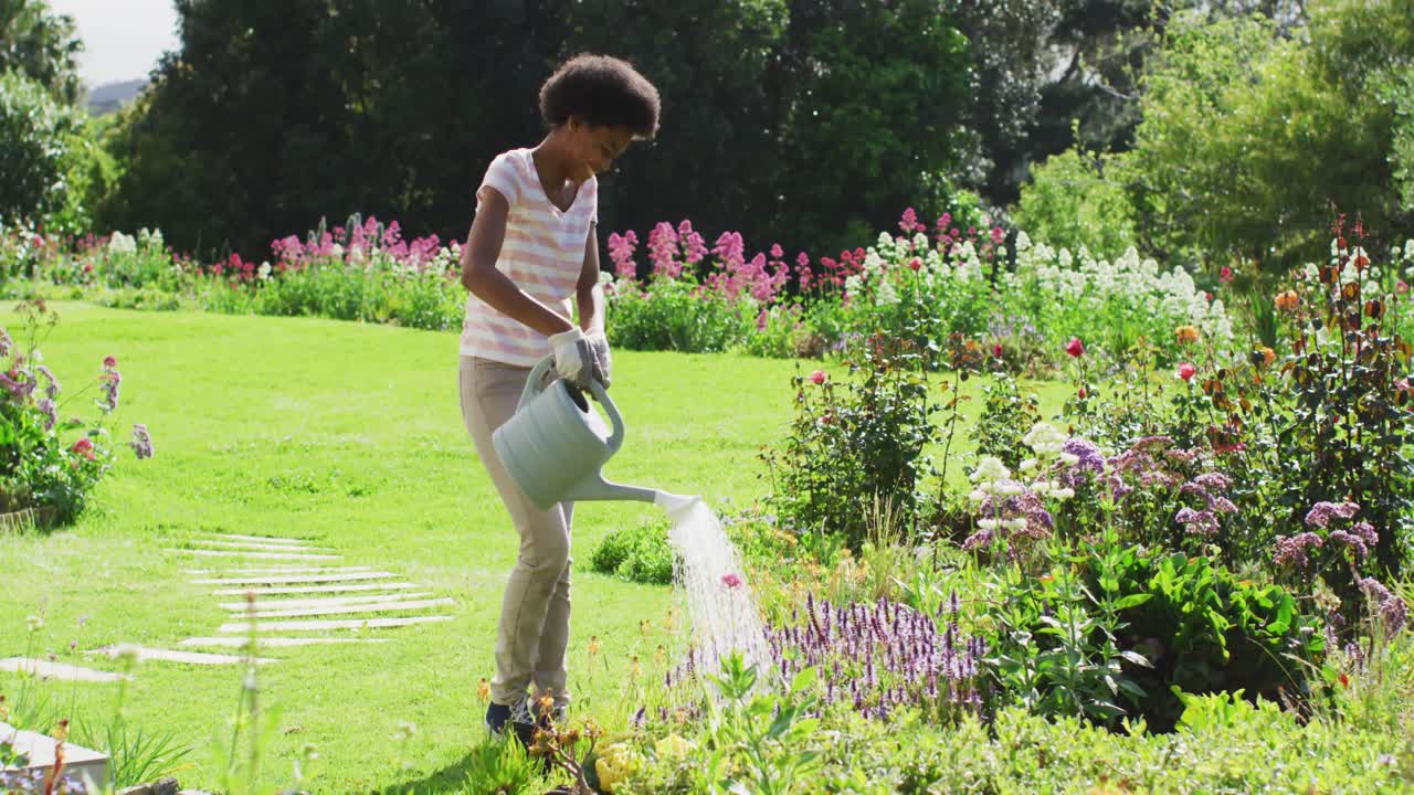 행복한 아프리카계 미국인 십대 소녀가  ⁇ 은 정원에서 식물에 물을 주고 있습니다.