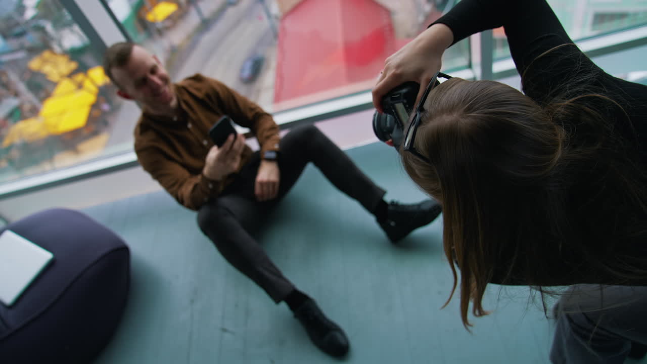 Photo session in office. Female photographer taking photos of businessman sitting on a floor with a mobile phone near the window with a city view.