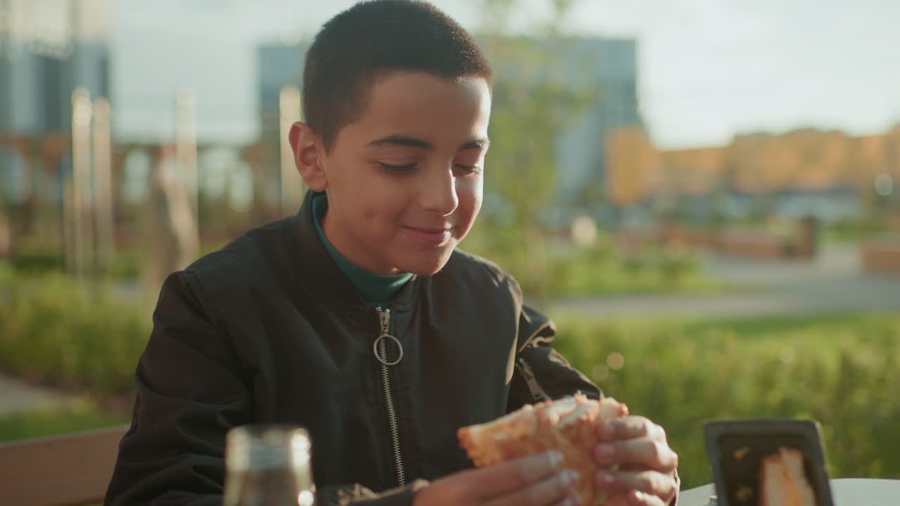 Happy boy sitting outdoors enjoying bite of pizza with juice bottle on wooden table, savoring meal with content expression under warm sunlight in casual park setting