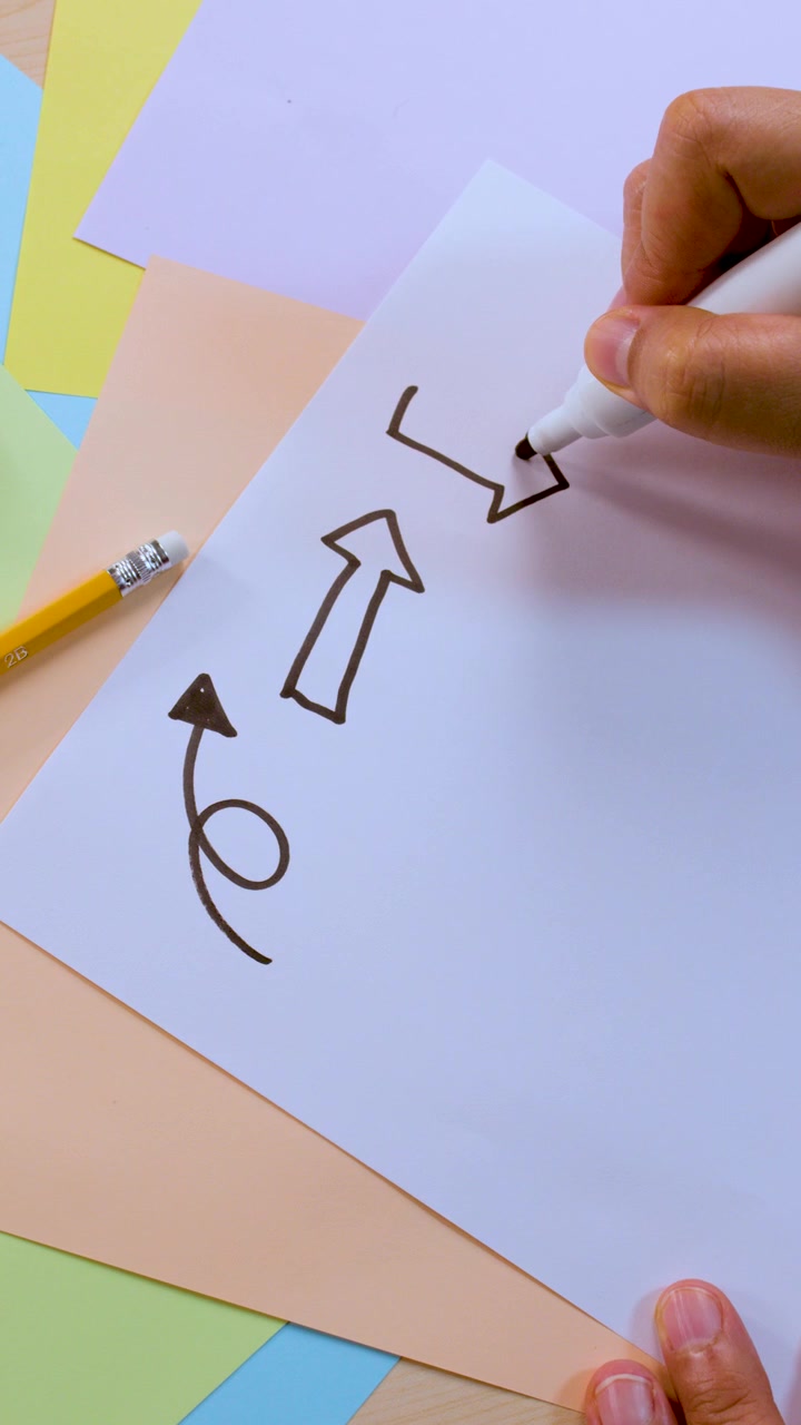 Una persona disegna varie frecce nere e linee curve su carta bianca in un ambiente luminoso e colorato