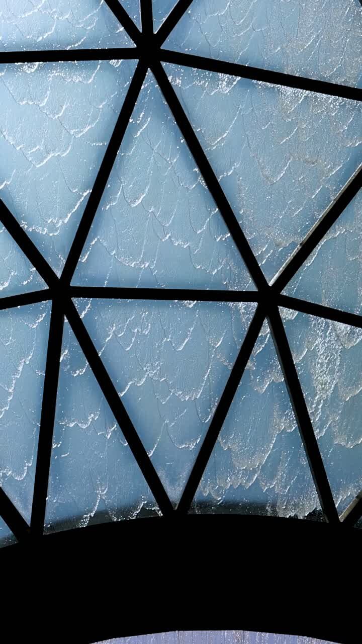 La telecamera si inclina verso l'alto sotto una cupola di vetro geometrica, rivelando una cascata interna e fogliame in silhouette in una luce diurna brillante e diffusa.
