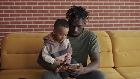 African-american-father-sitting-with-son-on-sofa-using-smartphone