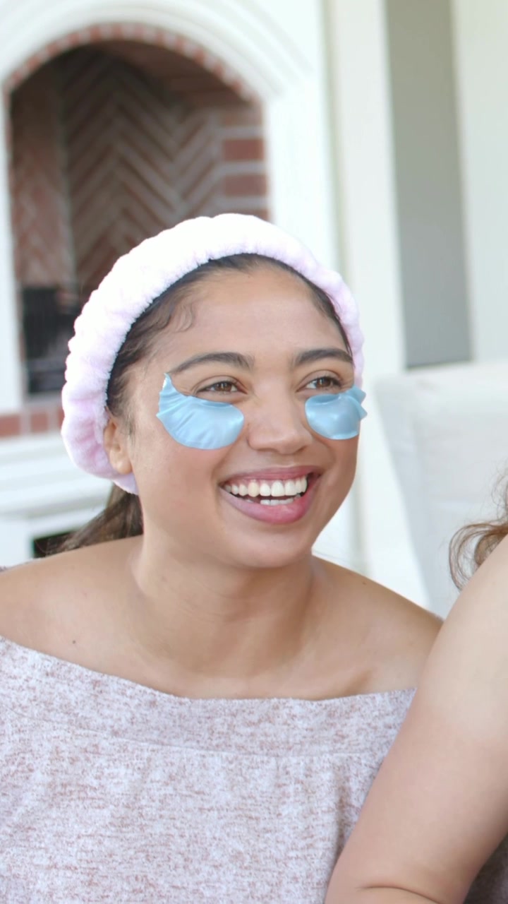 Vertical video: Smiling woman with pink headband blue eye gels leaning to friend on sofa after cue