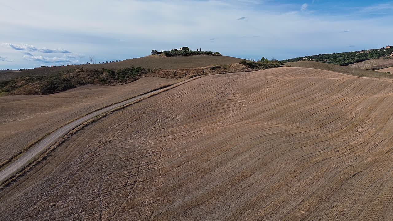 Panoramic view of Tuscany's golden rolling hills with a curving dirt path leading towards a hilltop villa under a blue sky.