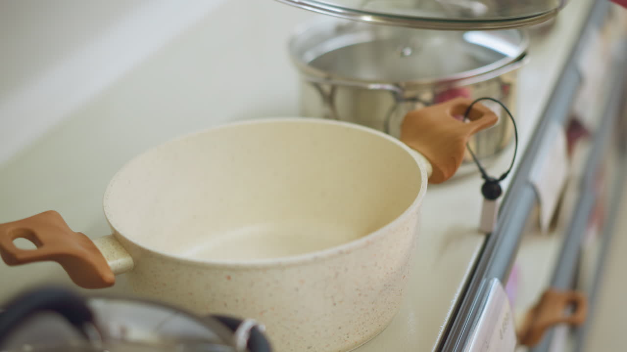 Hand of a lady in pink dress carefully opens a modern cookware pot with wooden handles in a kitchenware store, the pot has a speckled design, and it is part of a display with other pots