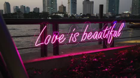 Eine-Atemberaubende-Leuchtreklame-Mit-Der-Aufschrift-„Love-Is-Beautiful“-Vor-Dem-Hintergrund-Der-Skyline-Und-Des-Flusses