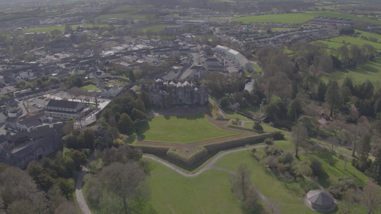An overhead shot from a drone that pans left around the front of Birr Castle in Birr, Co. Offaly that sits directly beside an Irish town. The castle is surrounded by houses, trees and Irish nature