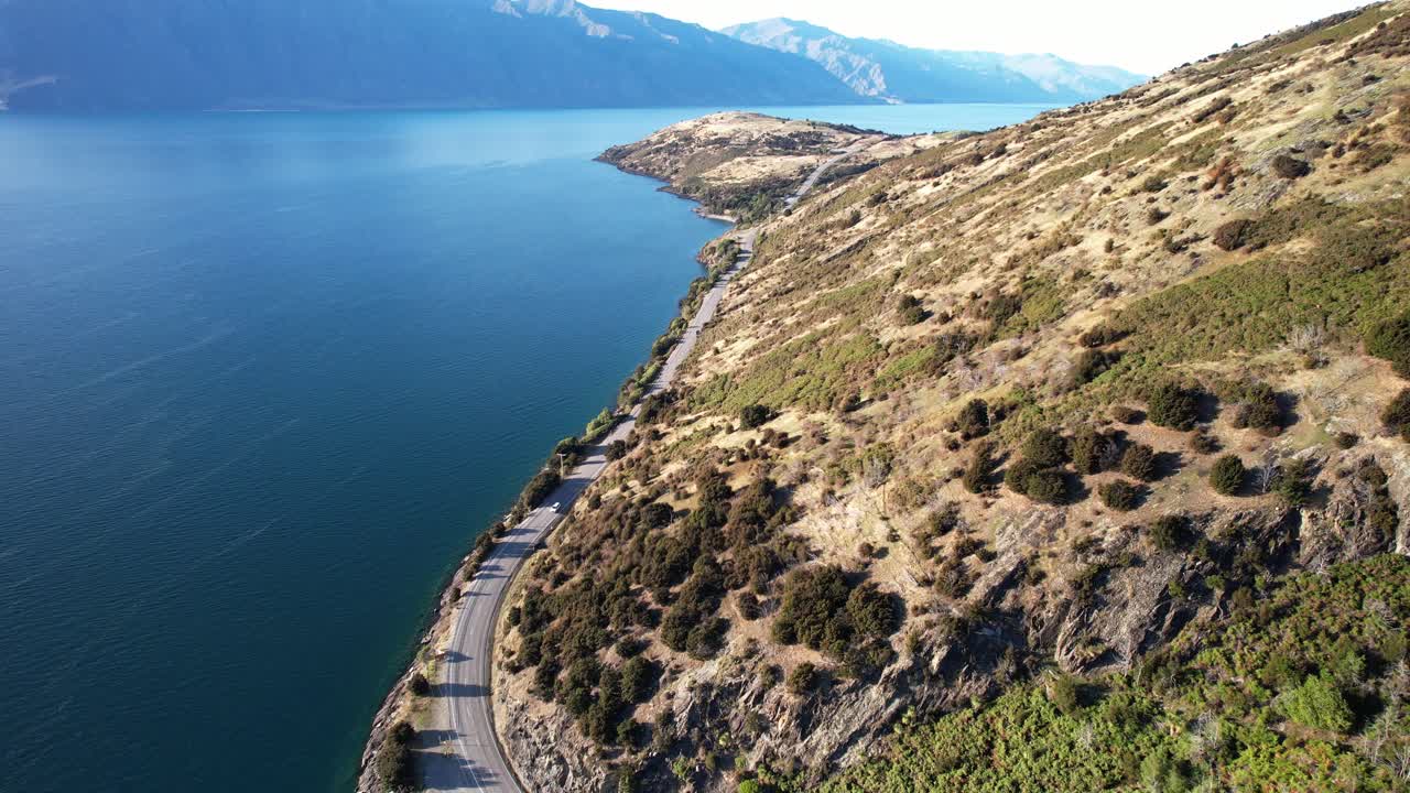 Road Along The Tranquil Blue Waters Of Lake Hawea In Summer In South Island, New Zealand. - aerial shot