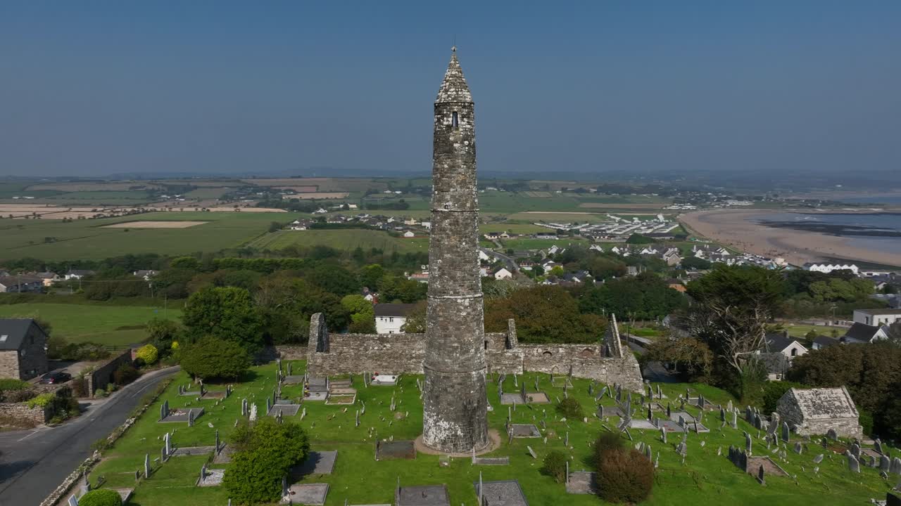St. Declan's Monastery, Ardmore, County Waterford, Ireland, September 2024. Drone orbits clockwise around weathered Round Tower with sweeping coastal views of the beach from the grassy hillside.