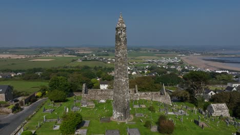 st. declan's monastery, ardmore, county waterford, ireland, september 2024. drone orbits clockwise around weathered round tower with sweeping coastal views of the beach from the grassy hillside.
