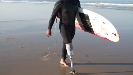 vertikale bewegung eines männlichen surfers mit künstlichem bein, der am strand entlang läuft und ein surfbrett unter dem arm hält 1