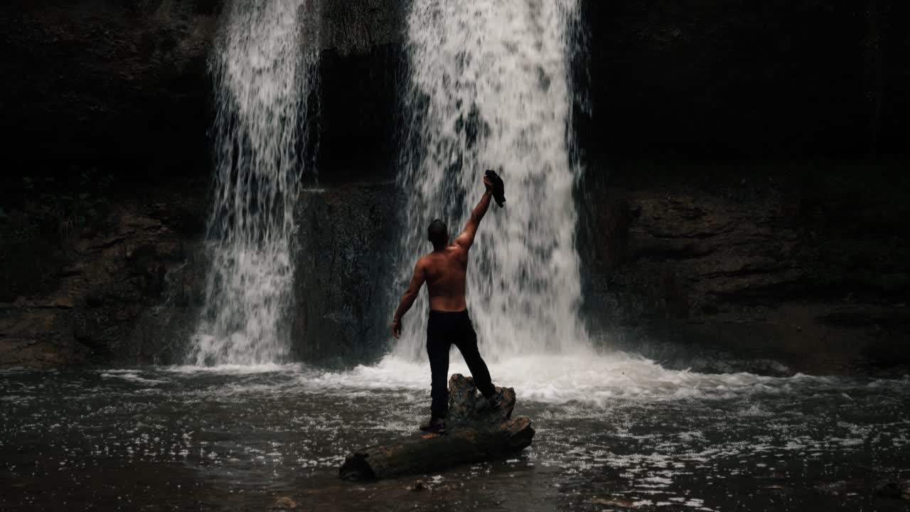 A person, a man, stands on a broken branch in front of a waterfall in a mystical forest. Shot in slow motion, upper body bare. His back is visible, as he raises his arms. Epic Landscape. Rainy day.