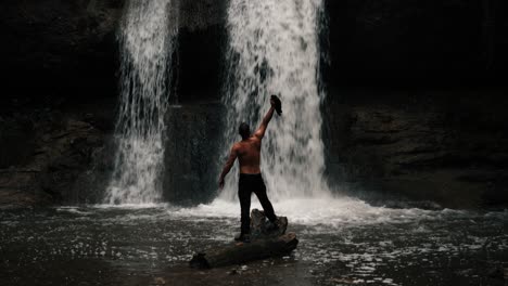 a person, a man, stands on a broken branch in front of a waterfall in a mystical forest. shot in slow motion, upper body bare. his back is visible, as he raises his arms. epic landscape. rainy day.