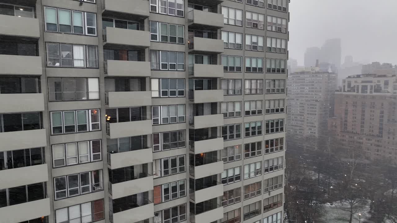 Vista aérea de una metrópolis estadounidense en el centro durante una tormenta de nieve invernal, torre residencial de gran altura con ventanas iluminadas, horizonte neblinoso de fondo, atmósfera urbana fría. Toma orbital