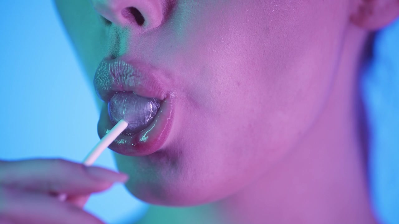 Macro in slow motion 4K di una donna che lecca un lecca-lecca rosa, primo piano cinematografico giocoso con sfondo blu vibrante, luce neon rosa e verde brillante sul suo viso, scena vibrante artistica e di tendenza