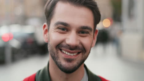 close-up view of young caucasian man with backpack looking aside in the street, then turns his face and smiles at the camera