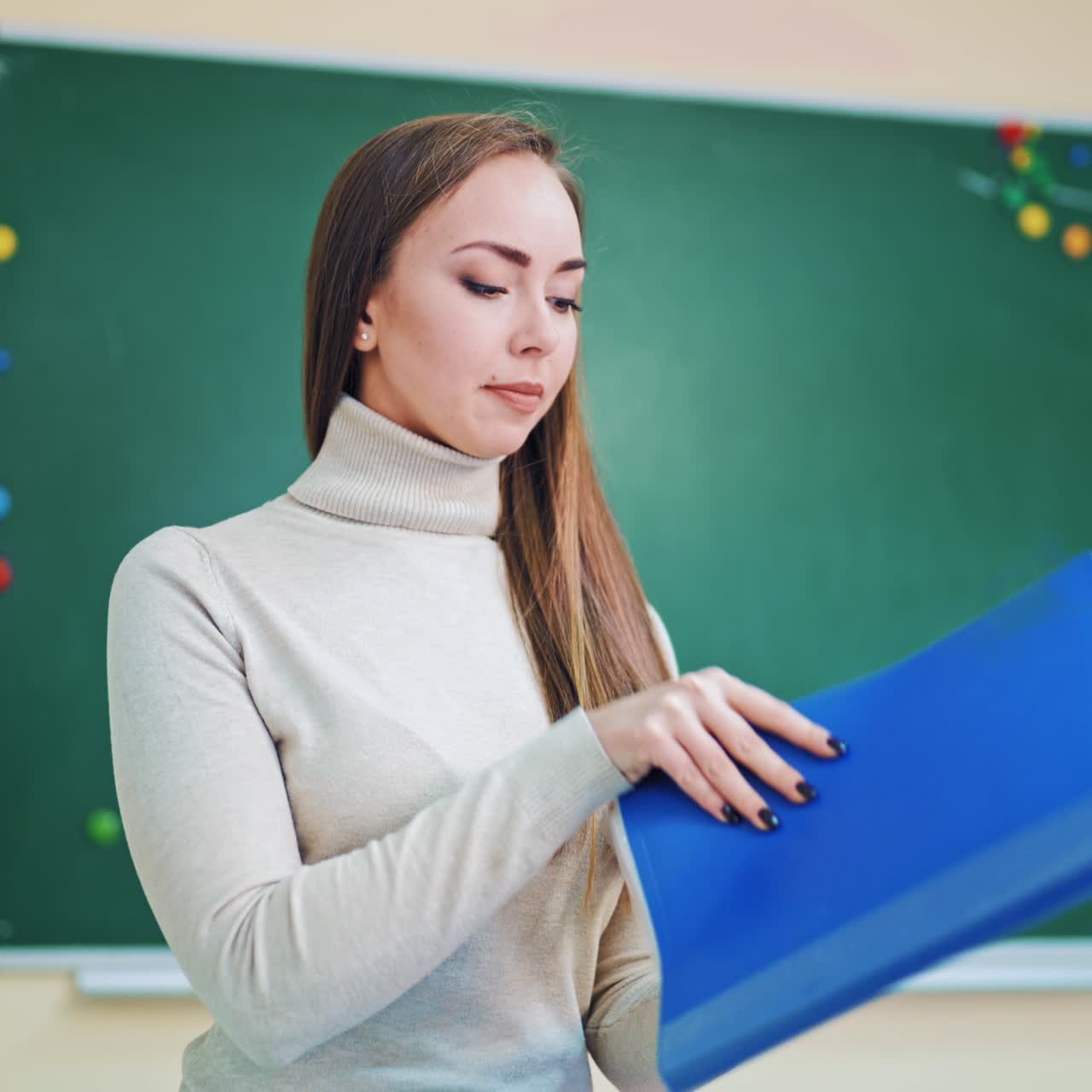 Beautiful teacher with a school register. Portrait of a young female educator looking into the folder on the blackboard background in the classroom.