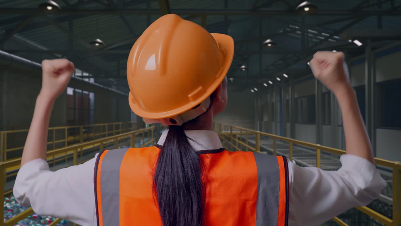 Primo piano, vista da dietro di un'ingegnere donna con elmetto di sicurezza che esulta con le mani alzate mentre lavora, ispezionando una fabbrica di riciclaggio della plastica con nastro trasportatore di bottiglie.