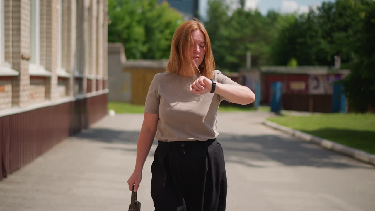 Woman checking wristwatch while walking outdoors under bright sunlight, expression showing concern about being late for meeting, holding handbag, wind gently moving hair