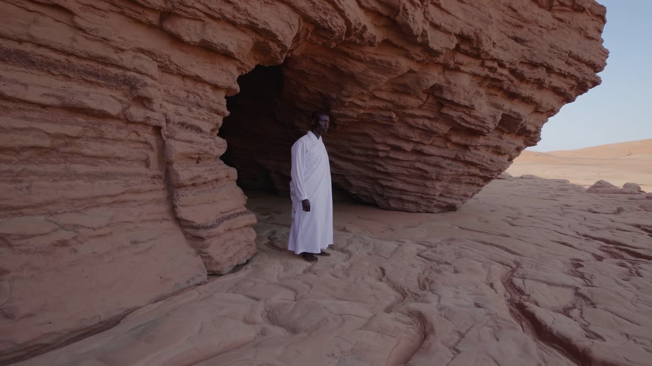 Monk wearing traditional white clothing standing in the shade of a large rock formation in a sunny desert, contemplating the vast expanse of sand and dunes