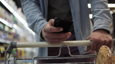 cropped close up footage of a young man's hands with stylish watches pushing trolley cart against blurred background and shopping in grocery supermarket. a guy using modern smartphone and choosing fresh food