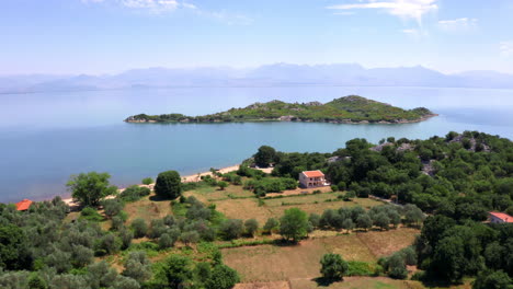 una pequeña isla con vegetación exuberante, aguas cristalinas alrededor, cerca de la orilla del lago skadar en montenegro con casas y botes en la playa, horizonte montañoso brumoso, toma aérea 4k