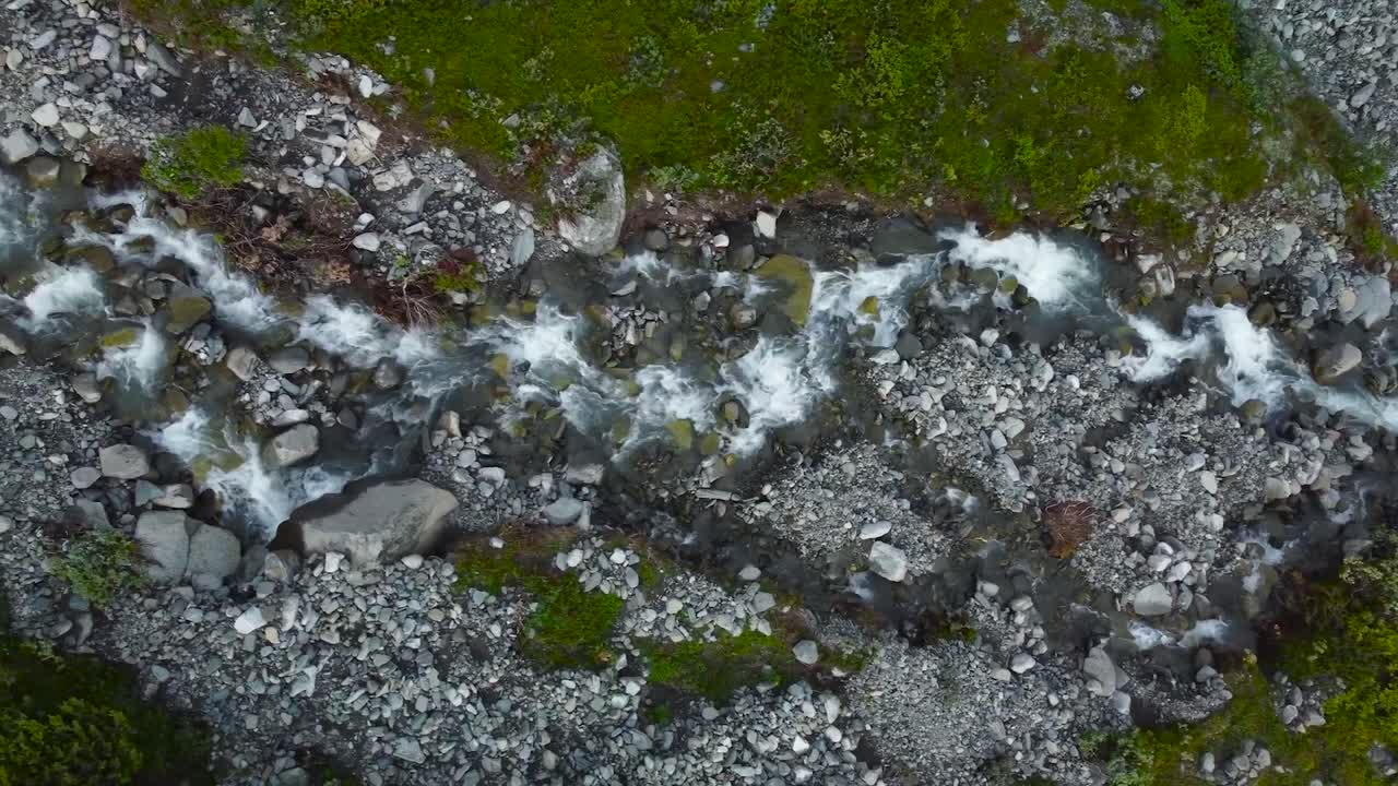 Top down aerial view flying over a narrow fast flowing river in Sweden during a cloudy day, The water is flowing through rocky gray terrain with green moss and short grass visible on the sides. Foamy.