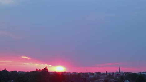 Lapso-De-Tiempo-De-La-Hermosa-Puesta-De-Sol-Roja-Escénica-Sobre-La-Ciudad-Con-El-Sol-Brillando-A-Través-De-Las-Nubes,-Plano-General