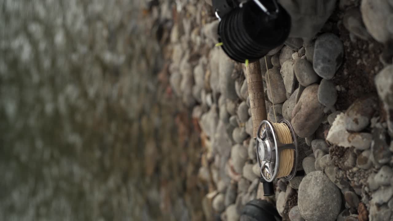 Formato verticale: primo piano di una mano che appoggia una canna da pesca a mosca su ghiaia e rocce di un fiume di montagna.