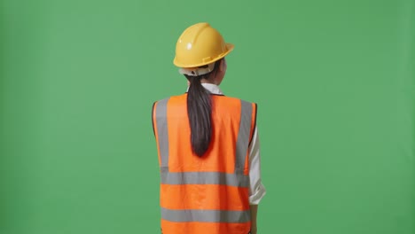 vista trasera de una ingeniera asiática con casco de seguridad hablando en un teléfono inteligente mientras está de pie en el estudio de fondo de la pantalla verde