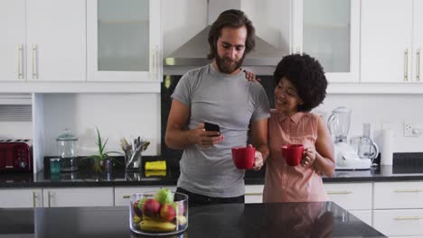 pareja de raza mixta sosteniendo una taza de café usando un teléfono inteligente en la cocina en casa