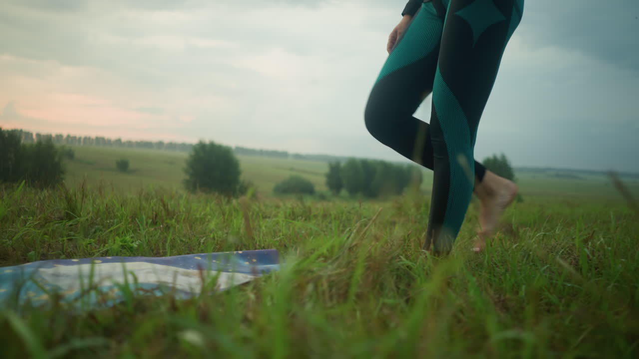 Leg view of a woman in black and green leggings walking barefoot on grassy field towards a mat on the ground, stepping onto the mat under a cloudy sky in a vast grassy field