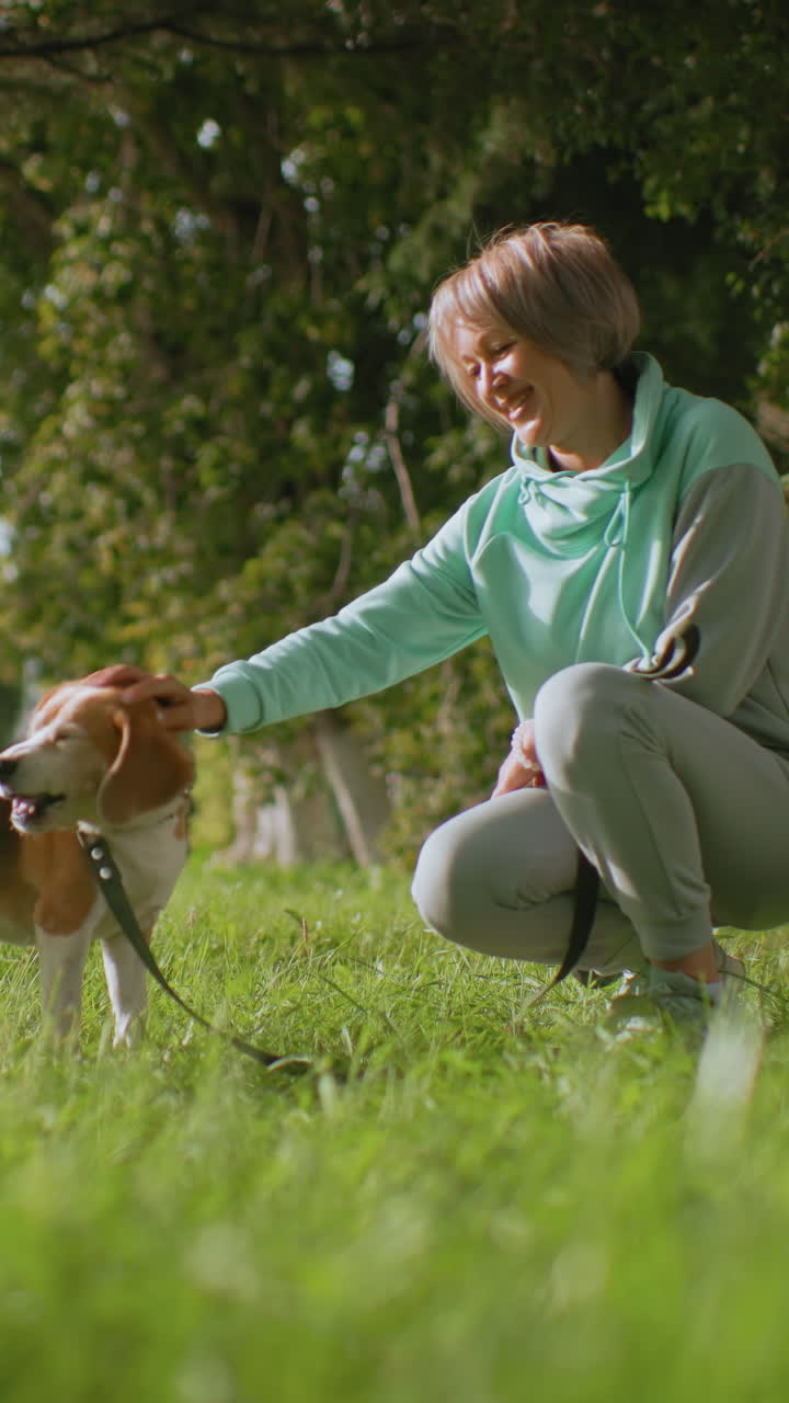 mujer asiática arrodillada ofreciendo una golosina a un beagle atado con correa en un parque iluminado por el sol, sesión de adiestramiento suave, b-roll de estilo de vida, luz natural cálida, hierba verde, dueña paciente sonriendo al perro, primer plano en ángulo bajo